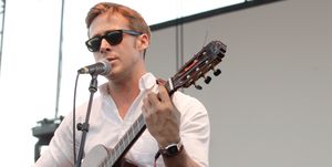 ryan gosling singing into a microphone and playing guitar on a stage, he wears sunglasses, a white collared shirt and gray pants with a belt