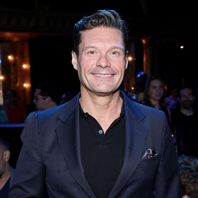 ryan seacrest smiles at the camera, he wears a navy blue blazer with a multi color pocket square over a black polo shirt