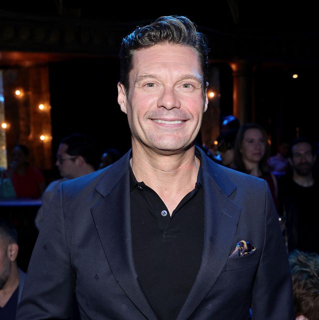ryan seacrest smiles at the camera, he wears a navy blue blazer with a multi color pocket square over a black polo shirt