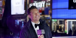 ryan seacrest wearing a tuxedo and jacket as he smiles off camera while holding a microphone