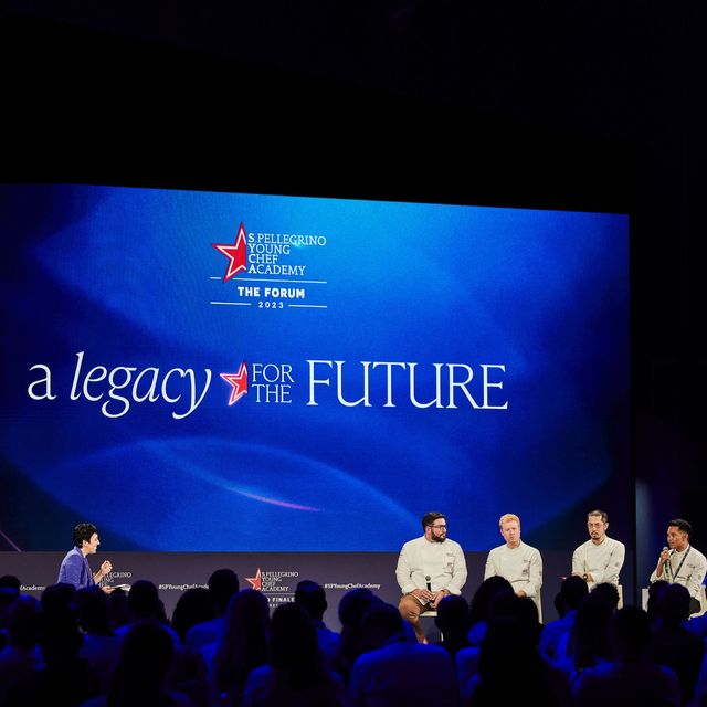 some of the competing chefs sit on stools on a stage, underneath a big blue screen with the worlds a legacy for the future
