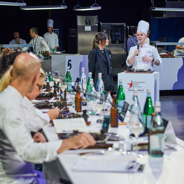 one of the competing chefs standing at a white block, presenting their dish and talking to judges sat at a long table in front
