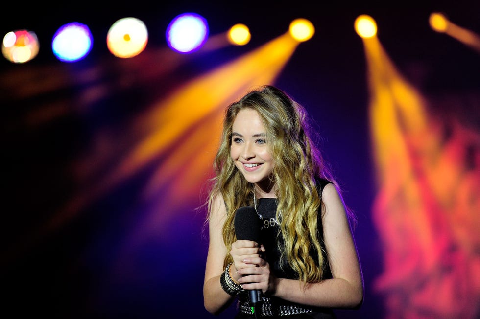 sabrina carpenter smiles, leans forward and looks left, she holds a wireless microphone with both hands and wears a black sleeveless outfit, multicolored stage lights stream in the background