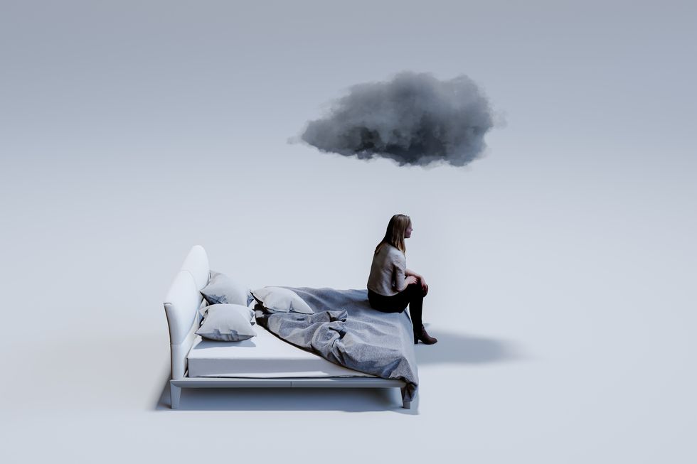 sad mid adult woman sitting on corner of double bed with dark cloud above head