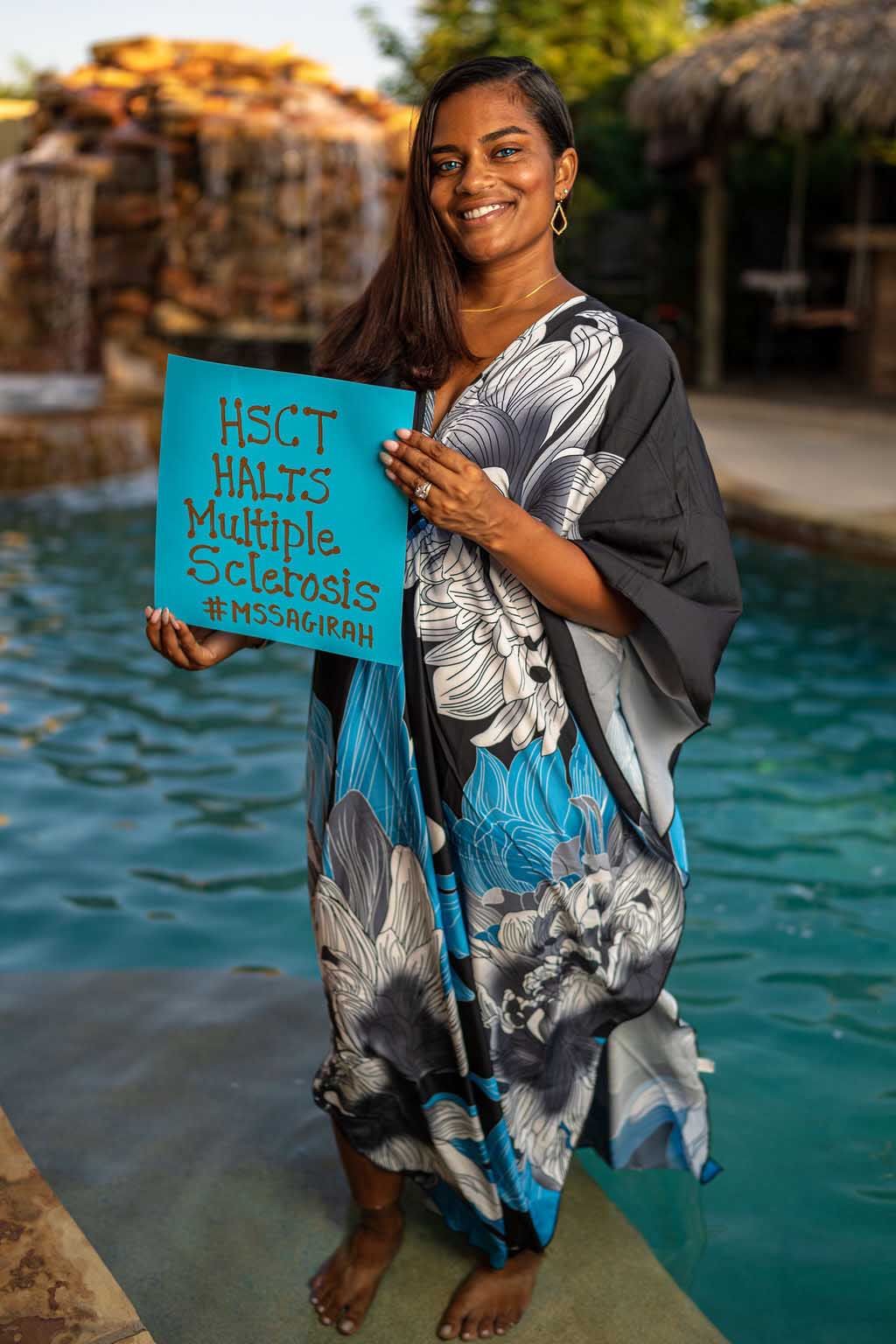 sagirah norris standing in front of a pool smiling wearing a long blue black and white dress, she carries a sign that lists her invisible disabilities