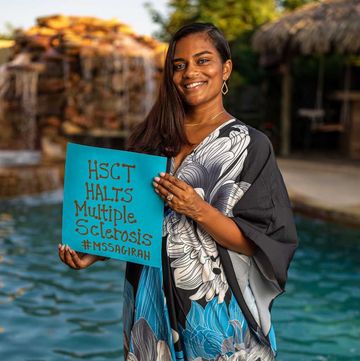 sagirah norris standing in front of a pool smiling wearing a long blue black and white dress, she carries a sign that lists her invisible disabilities