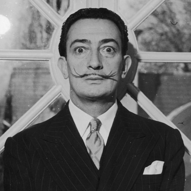 salvador dali stares wide eyed into the camera, he has his signature thin mustache and wears a suit with a pocket square