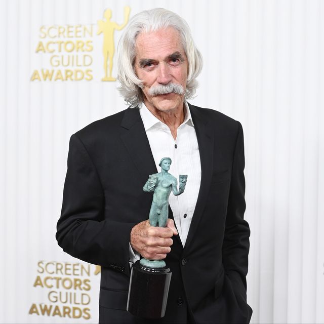 sam elliott holds a bronze statue from the screen actors guild award and looks at the camera, he wears a black suit jacket and white collared shirt that is unbuttoned at the neck
