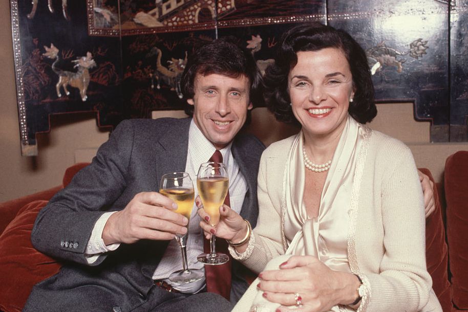 richard blum and dianne feinstein smile for a photo while sitting on a red couch and holding champagne flutes, he wears a gray suit with a white shirt and red tie, she wears a white sweater and blouse with pearls