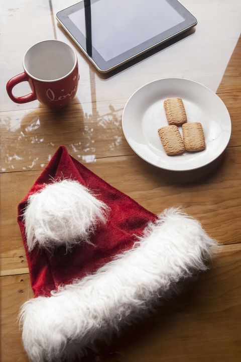 a santa hat on a wood table with a plate of cookies, a mug, and a tablet