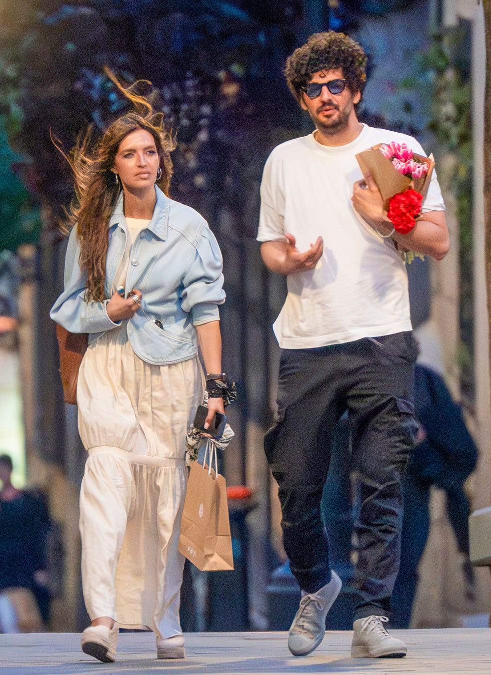 Una pareja caminando por una calle cargando bolsas y flores.