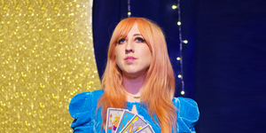 sarah potter, a white woman with long pink hair and bangs, holds four tarot cards in her hand she's wearing a blue velvet dress and looking upwards
