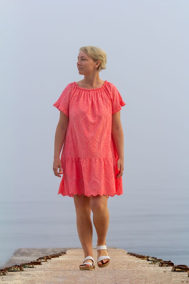 a satisfied middle aged woman in a pink sundress walks by the sea, looking away. female happiness and wellbeing