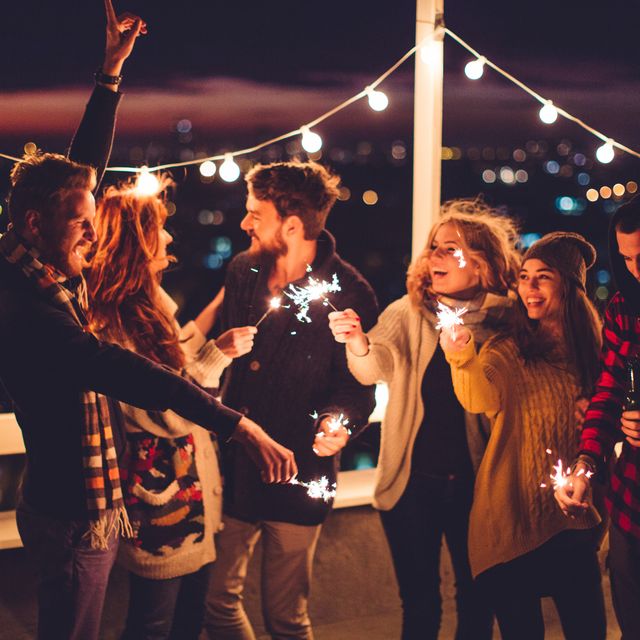 couple of friends having a party on the rooftop of the building at sunset drinking bottles of beer and holding sparklers wearing knitted sweaters, hats and scarfs they are happy and joyful