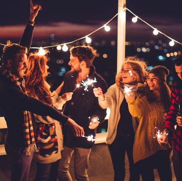 couple of friends having a party on the rooftop of the building at sunset drinking bottles of beer and holding sparklers wearing knitted sweaters, hats and scarfs they are happy and joyful