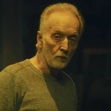 tobin bell as jigsaw in saw x, an older man with grey hair and beard stands in a dark room looking at the camera, he wears a long sleeve grey top