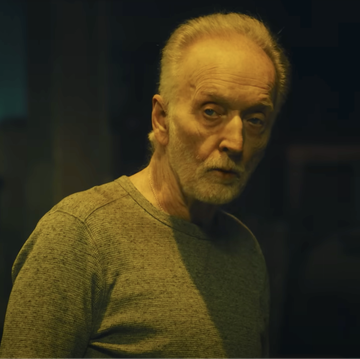 tobin bell as jigsaw in saw x, an older man with grey hair and beard stands in a dark room looking at the camera, he wears a long sleeve grey top