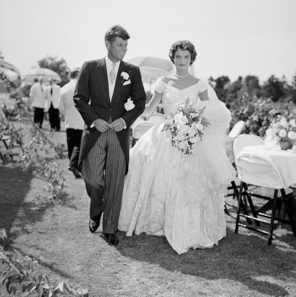 john f kennedy and jackie kennedy walk arm in arm on grass, he wears a suit, she wears a large wedding dress and carries a floral bouquet