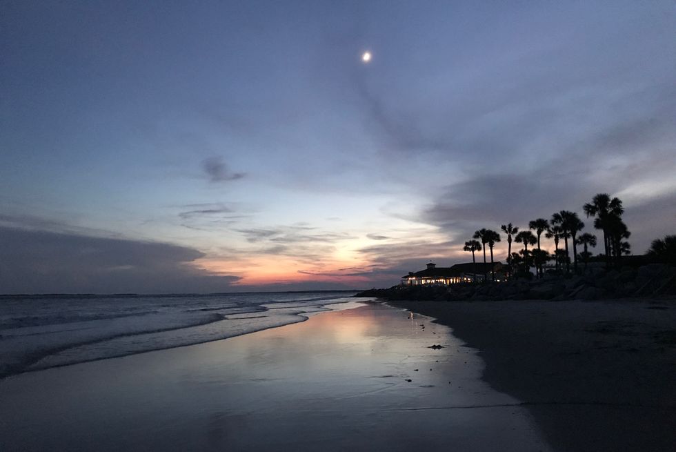 scenic view of beach during sunset with moon, outline of palm trees and lighted structure in the distance