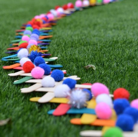 colored craft sticks with pom poms on top are lined up on grass as part of a science experiments for kids about chain reactions and potential and kinetic energy