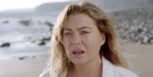 ellen pompeo, a white actress with brunette hair, stands on a beach in a white button down shirt, looking into the distance