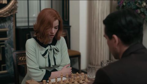 anya taylor joy starring in the queen's gambit sitting at a chess table wearing a green long sleeve shift dress with dark green panels and bow detail at the neck
