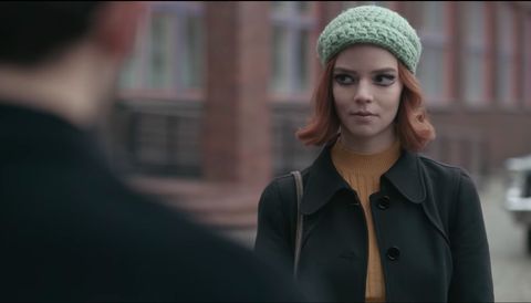 anya taylor joy starring in the queen's gambit, wearing a dark green wool coat with a pale orange high neck ribbed top and green knitted hat