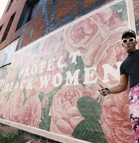 a man stands in front of a large mural that says "protect black women" over a floral background