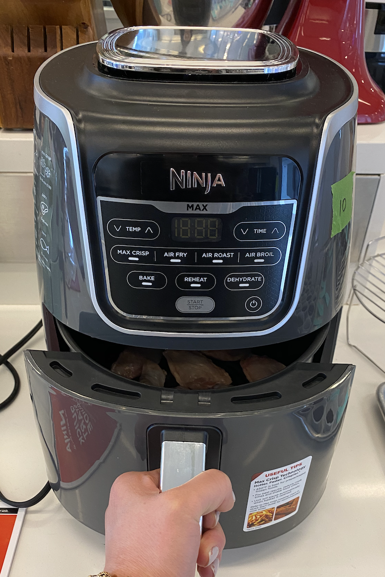 a hand opening the basket of the ninja air fryer max xl on a white counter in the good housekeeping institute