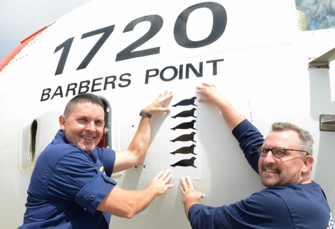 coast guard commander and noaa employee hold seven hawaiian monk seal decals in front of an hc 130 hercules airplane