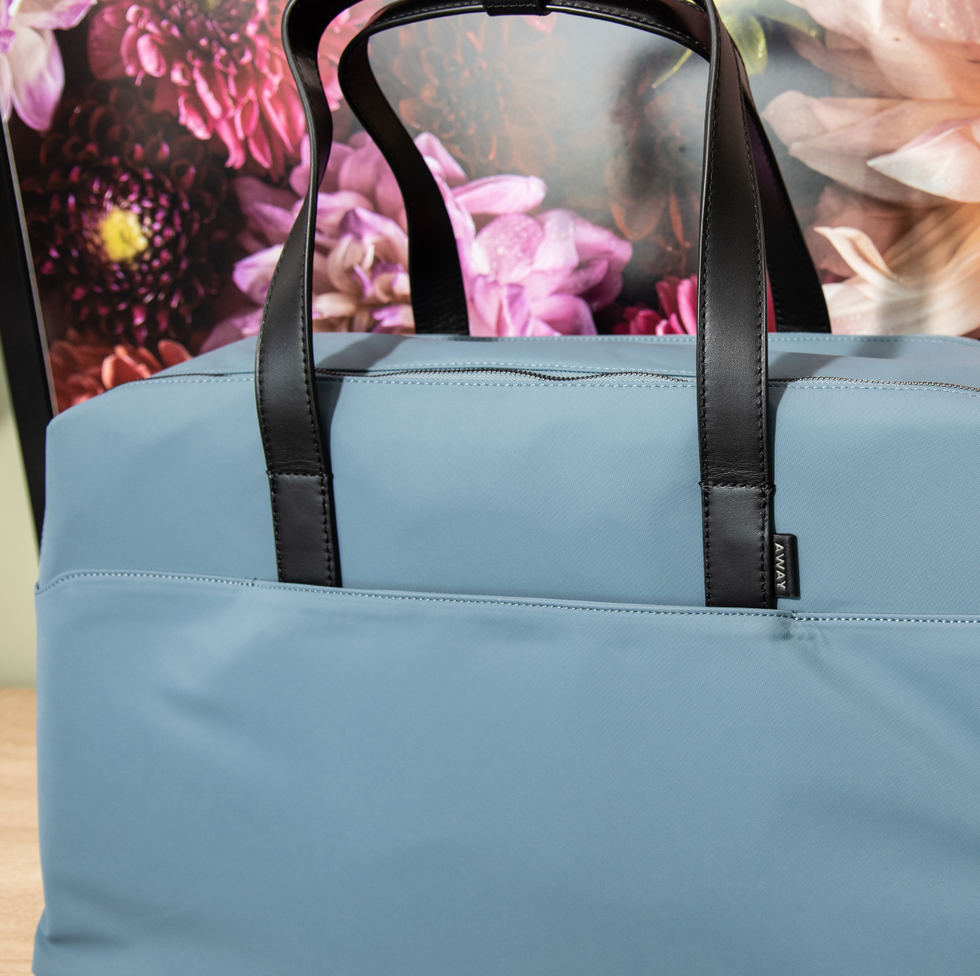 a blue away everywhere large bag in front of a floral backdrop, good housekeeping's best weekender bags
