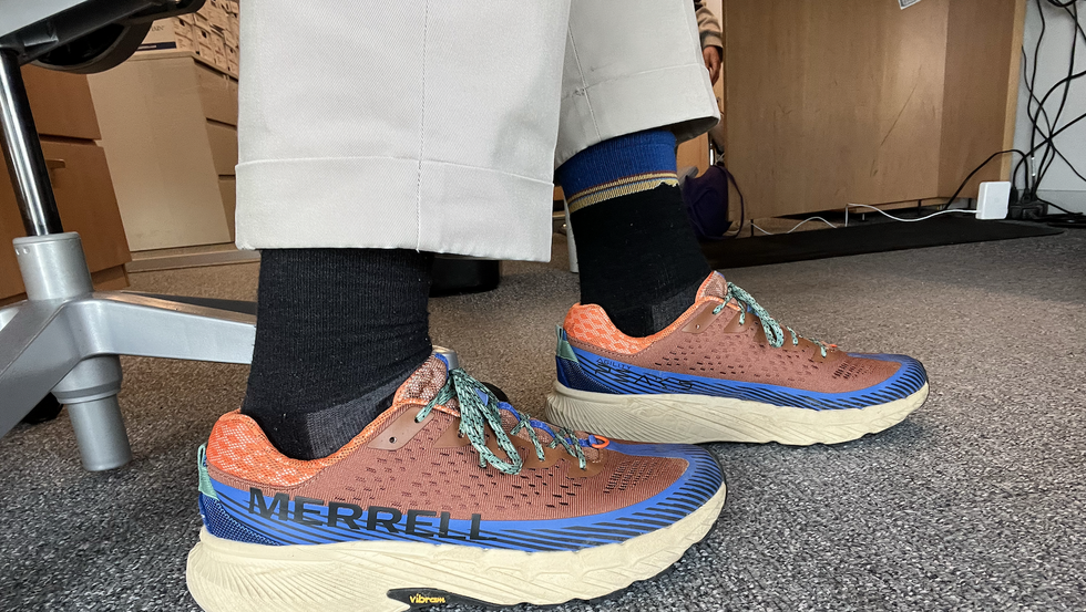 a tester wearing a pair of brown and orange merrell agility peak 5 sneakers in an office as part of good housekeeping's testing for the best walking shoes for men