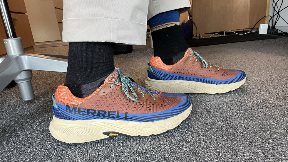a tester wearing a pair of brown and orange merrell agility peak 5 sneakers in an office as part of good housekeeping's testing for the best walking shoes for men