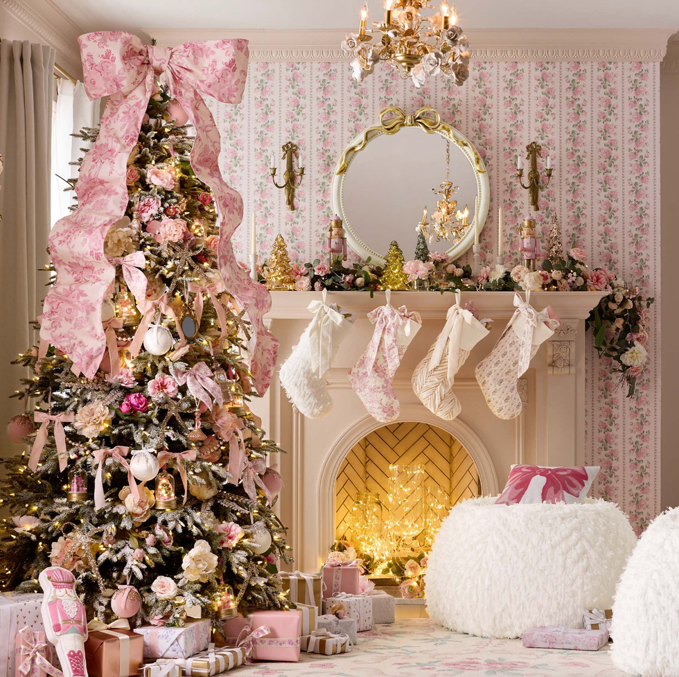 a beautifully decorated christmas tree stands in the corner, adorned with pink ribbons, ornaments, and floral accents nearby, a cozy fireplace features white stockings hanging from the mantle, along with decorative items and small evergreen trees the room has floral wallpaper and soft, inviting furnishings, including fluffy poufs presents are scattered on the floor, enhancing the festive atmosphere