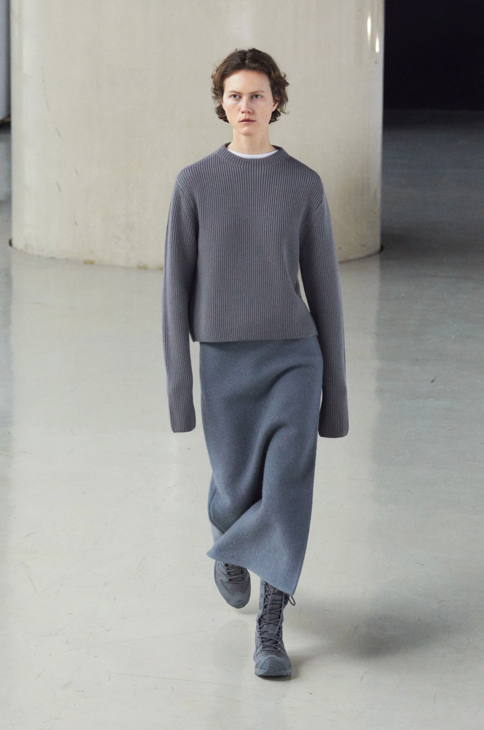 Model walking in a modern spacious environment wearing a gray ribbed sweater and a matching long skirt with gray boots