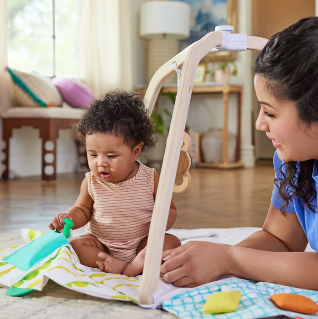 a baby plays on a play gym a mom and baby interact with a baby play gym, part of a good housekeeping list of the best play gyms