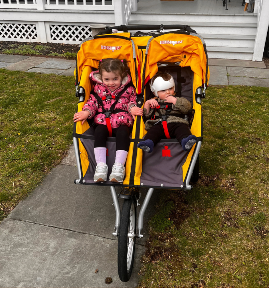 two siblings sitting in a yellow double jogging stroller in front of a house bob gear revolution flex 3 duallie double jogging stroller