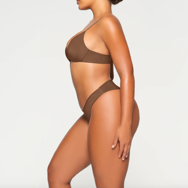 a side view of a woman modeling a brown bra from skims against a grey background