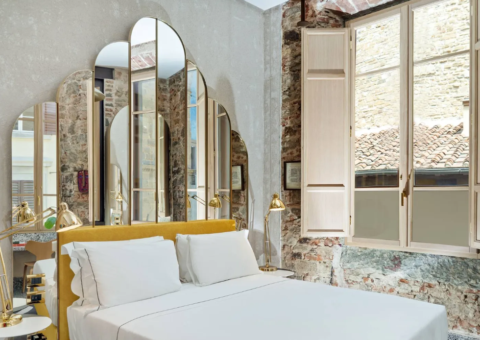 hotel room interior with mirrored panel behind headboard, open windows, exposed brick on a wall, and crisp white bedding