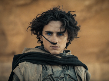 a figure with curly hair wearing a dark cloak against a sandy background