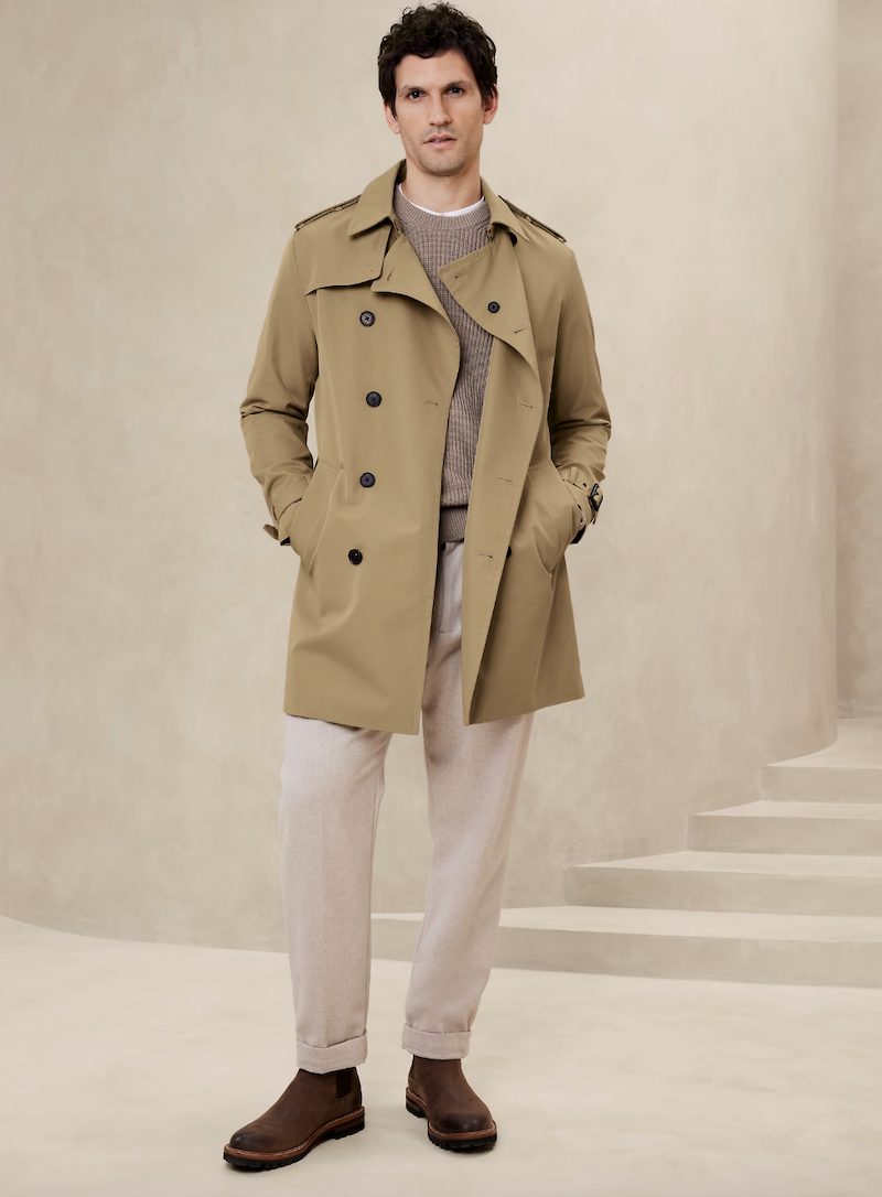 a man dressed in a tan trench coat over a gray sweater paired with lightcolored trousers and brown boots