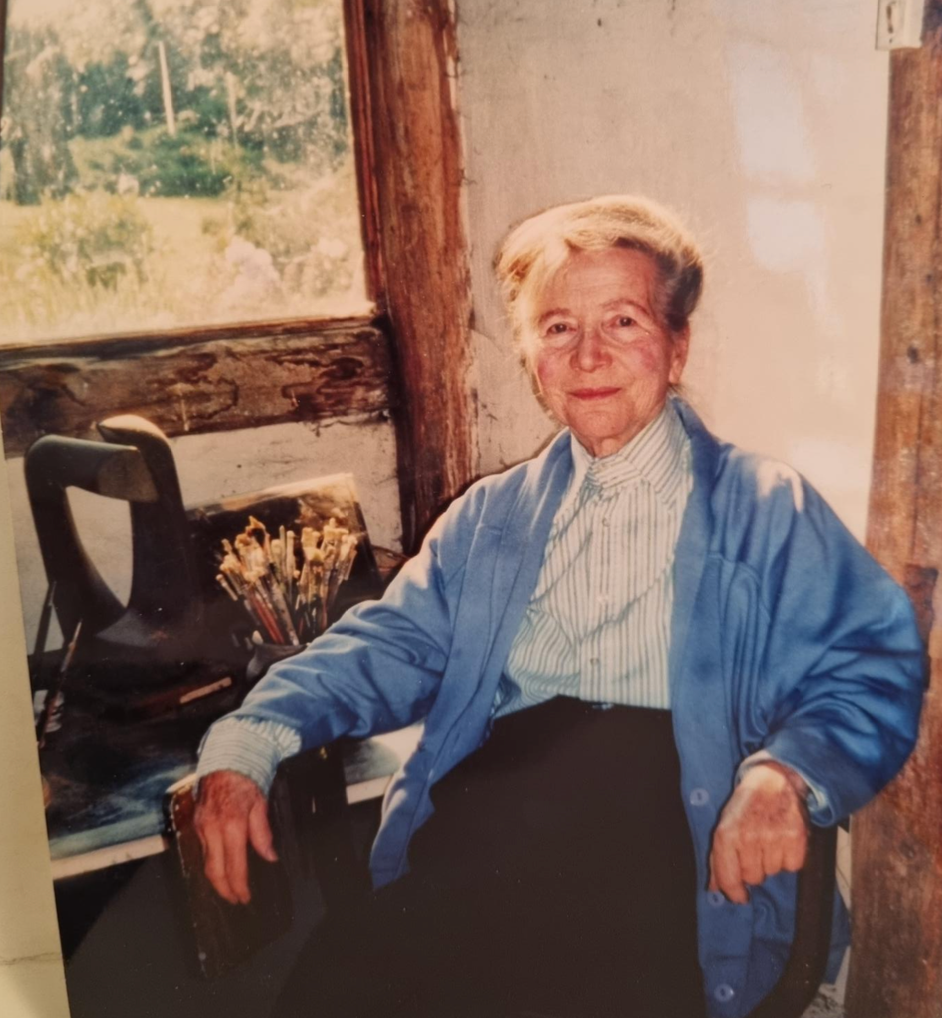 an elder person seated in a rustic interior surrounded by art supplies