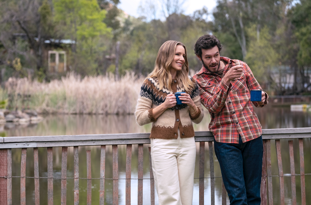 two individuals standing on a wooden railing by a lake holding blue mugs