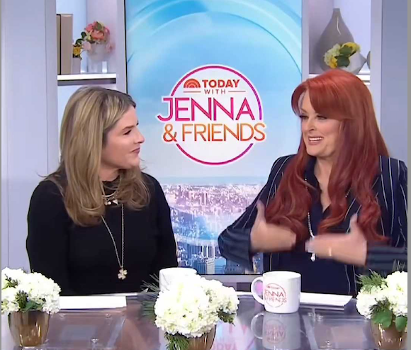 two women engaging in conversation on a talk show set with a floral centerpiece