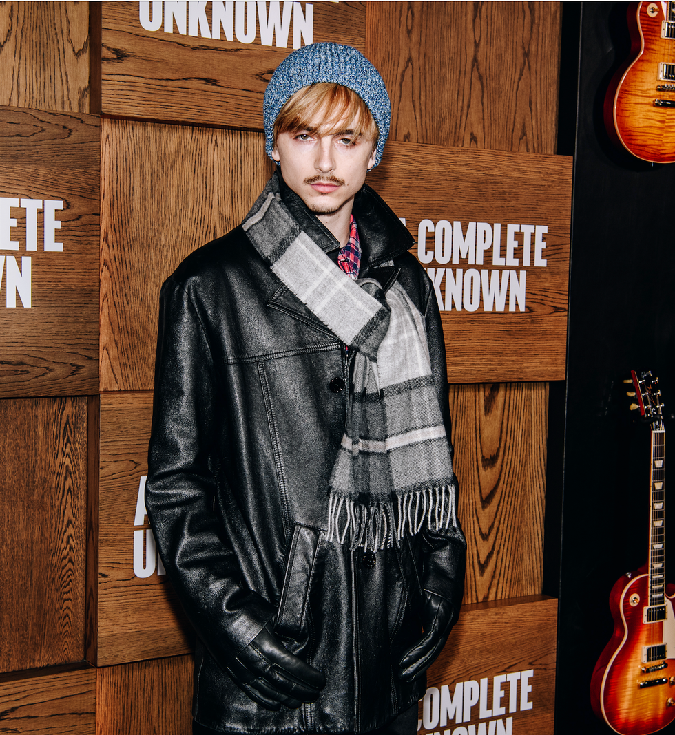 an elegantly dressed mannequin wearing a black leather jacket scarf and gloves next to guitars and a wooden wall