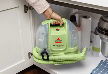 a person carrying the bissell little green mini portable carpet cleaner and storing it in a cabinet under the kitchen sink