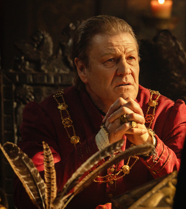 a seated figure with ornate clothing and jewelry hands clasped with a contemplative expression