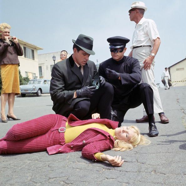 a woman lies on pavement as two men crouch beside her and three people stand in the background