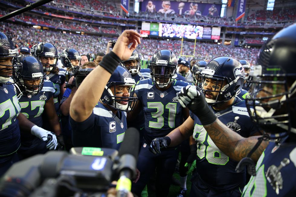 a group of seattle seahawks football players gathered in a huddle as quarterback russell wilson speaks from the center of them, with his hand raised in the air, during super bowl xlix