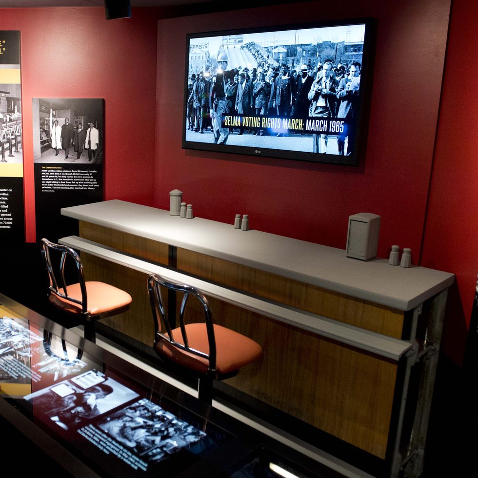 a woman looks at museum exhibit consisting with black displays of text and a television mounted on a red wall, and a diner counter with chairs underneath the television